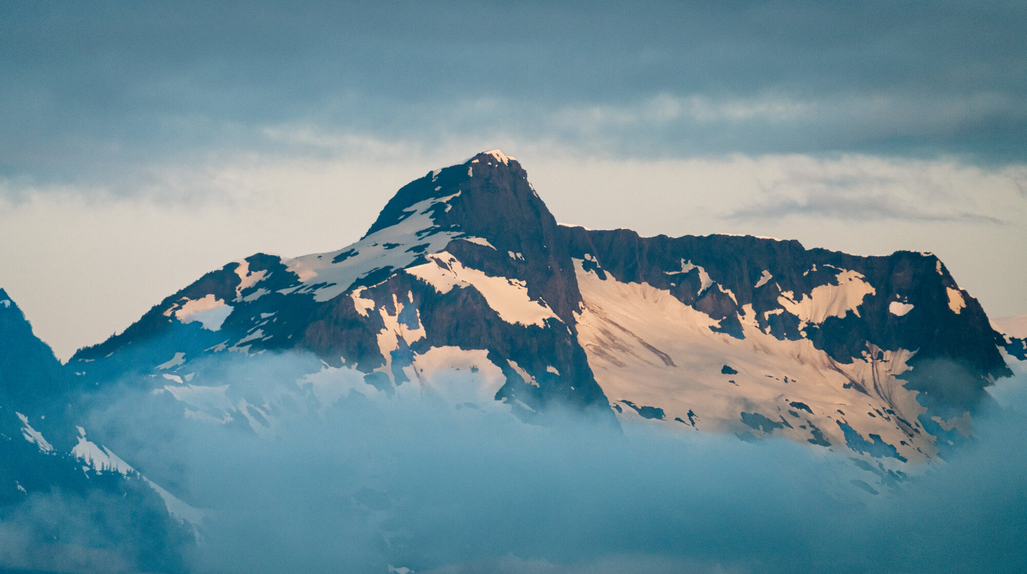 Broad snow-streaked coastal mountain rising through low gray clouds in Southeast Alaska