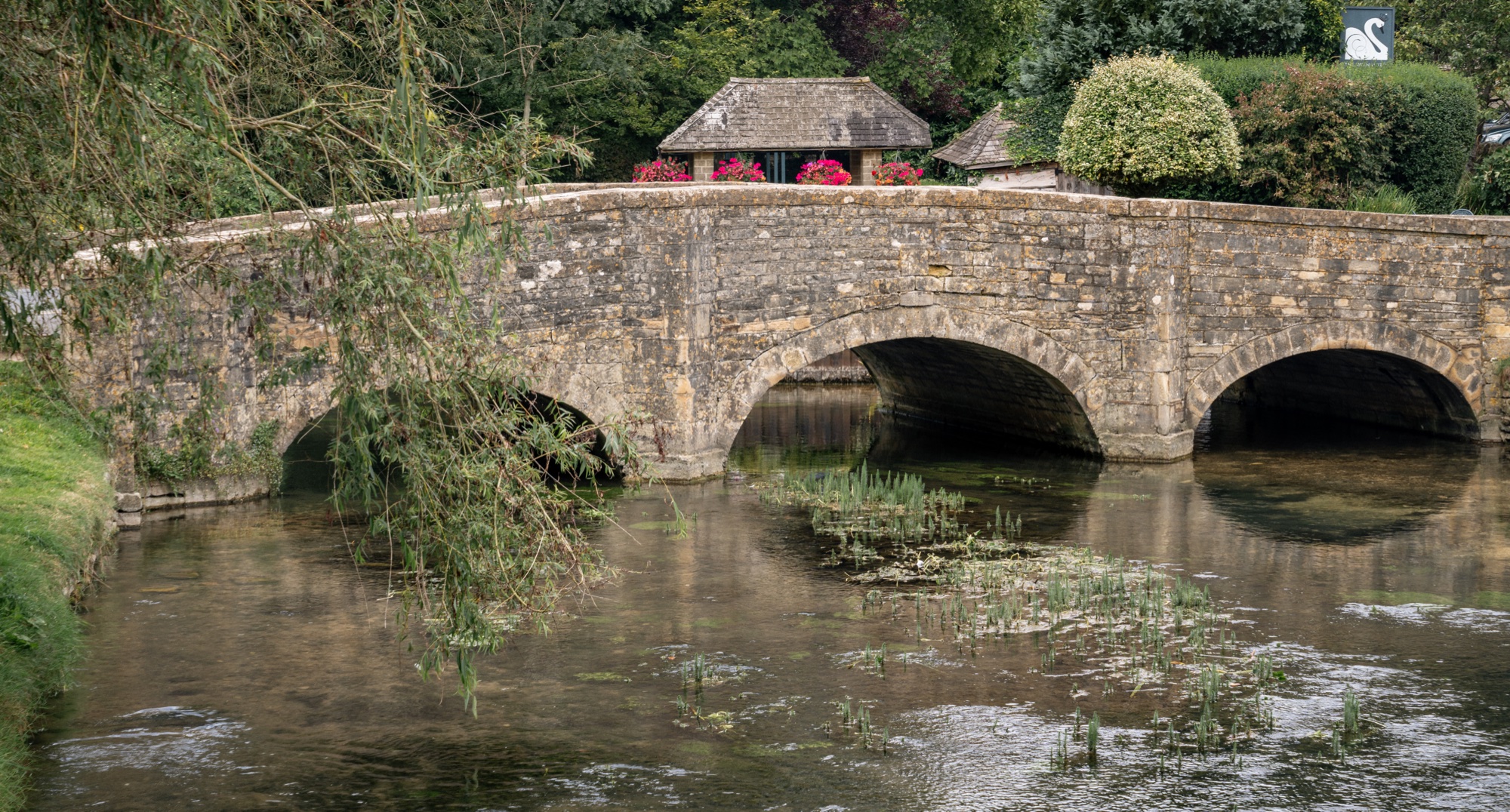 Low twin-arch Cotswold stone bridge crossing a clear river bordered by gardens