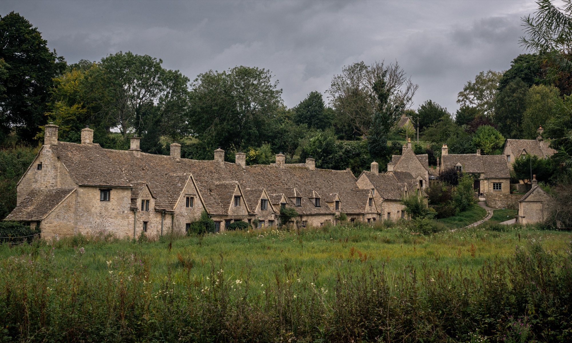 Long row of Cotswold stone cottages beyond a green meadow under a dark gray sky