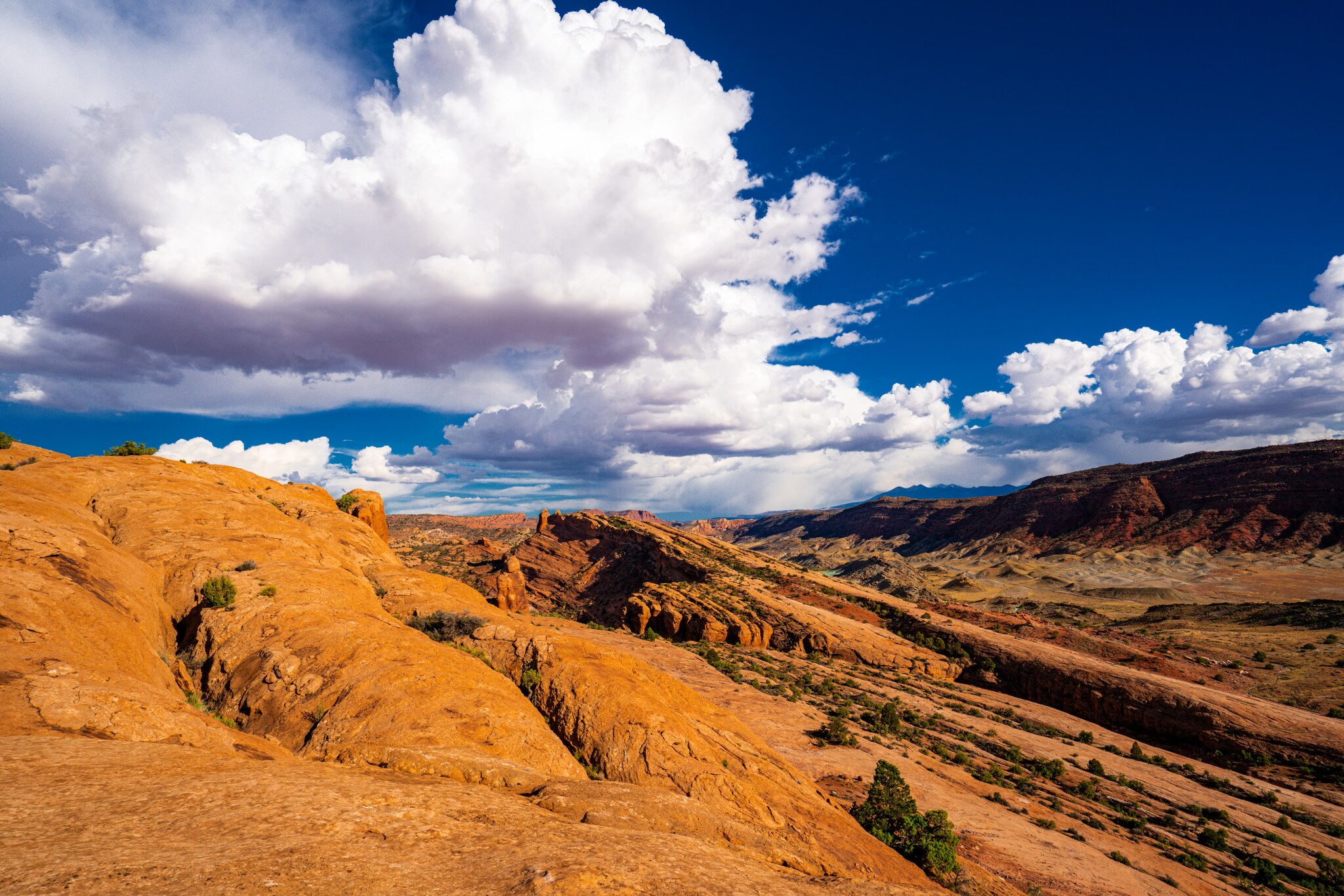 Golden slickrock descending into a broad Utah valley beneath towering white storm clouds and deep blue sky
