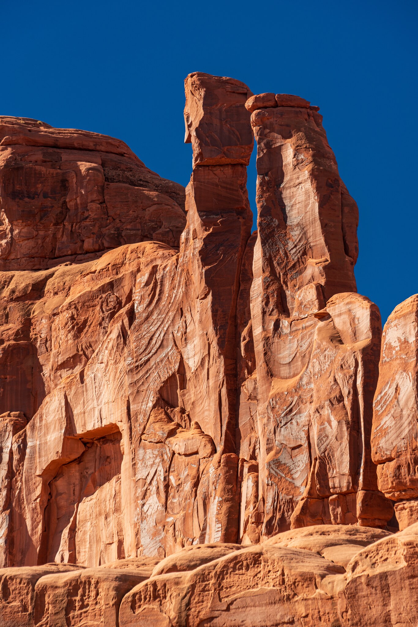 Two tall weathered sandstone towers rising beside a red cliff under a cloudless blue Utah sky